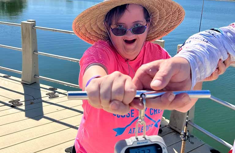 Woman smiling wearing a hat and sunglasses and holding up a fishing scale.