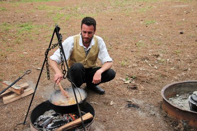 Person in period costume stirring something in a Dutch oven that is suspended over a campfire