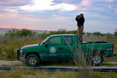 Game warden standing in bed of truck looking through binoculars