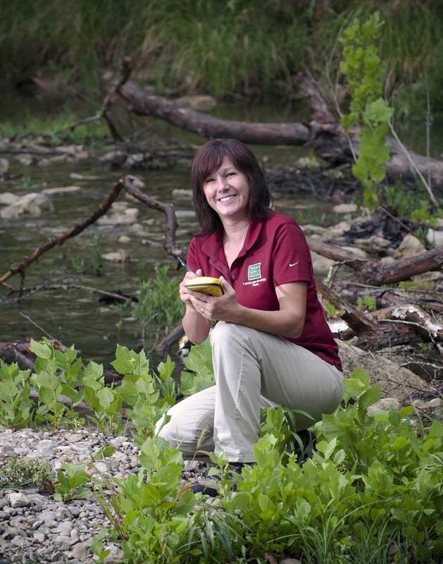 A female stream ecologist kneeling in a stream studying aquatic animal and plant life and doing research studies.