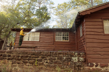 Firefighter standing on picnic table spraying roof of CCC cabin