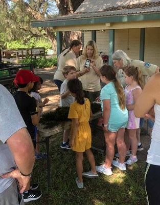 Group of kids on a field trip looking at items on a table as adults explain what they see.