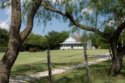 Farmhouse framed by mesquite trees and barbed wire fence.
