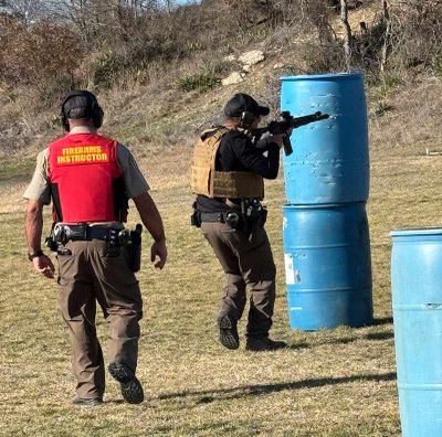 Man firing a rifle is followed closely by a firearms instructor.