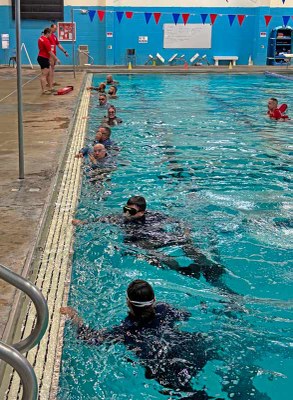 Trainees itouching the edge of an indoor swimming pool as lifeguards stand watch.