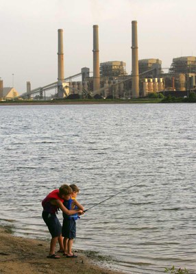 Two kids fishing on shore of lake with power plant across the water