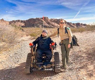 Ranger stands next to man sitting in a trackchair out on the trail.